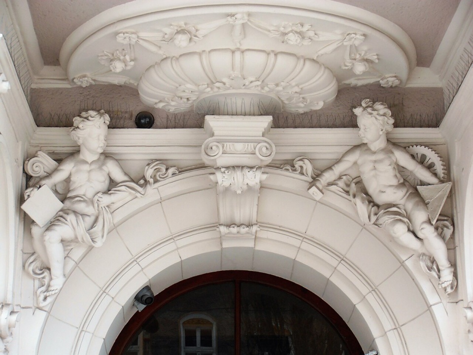 Putto z młotkiem, kielnią i kołem zębatym jako alegoria rzemiosła (po prawej stronie). Kamienica przy ulicy Śląskiej 38 w Szczecinie. Fot. Szczecinolog [wikipedia.org]